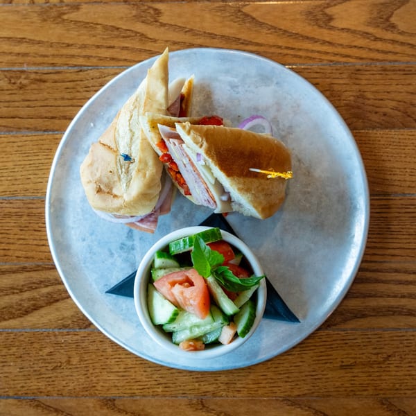 Two halves of a deli sandwich with a side of cucumber and tomato salad on a metal plate.