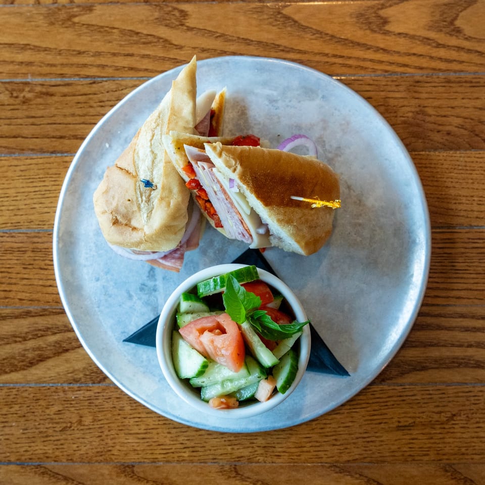 Two halves of a deli sandwich with a side of cucumber and tomato salad on a metal plate.