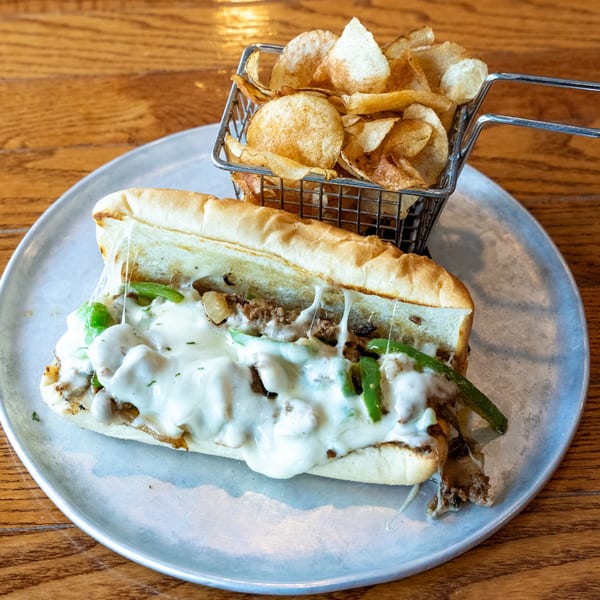 Cheesesteak sandwich with melted cheese, green peppers, and onions, served with a side of chips.