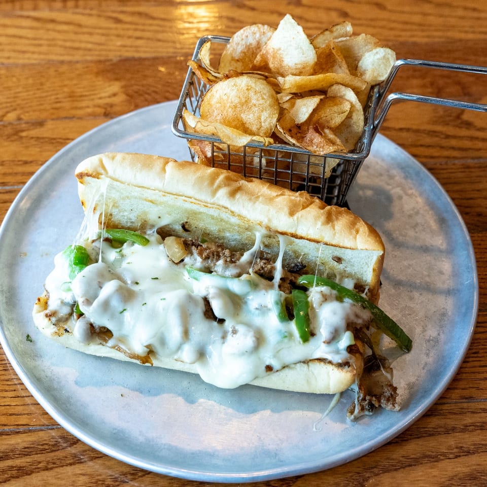 Cheesesteak sandwich with melted cheese, green peppers, and onions, served with a side of chips.