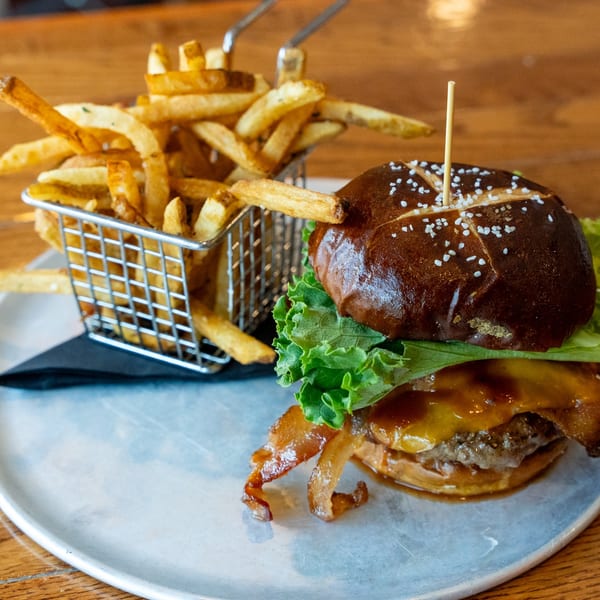 A gourmet burger with bacon, cheese, lettuce, and a pretzel bun, served with a side of fries.