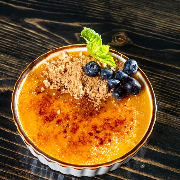 Crème brûlée dessert in a ramekin, topped with blueberries, mint, and crumble on a dark wooden table.