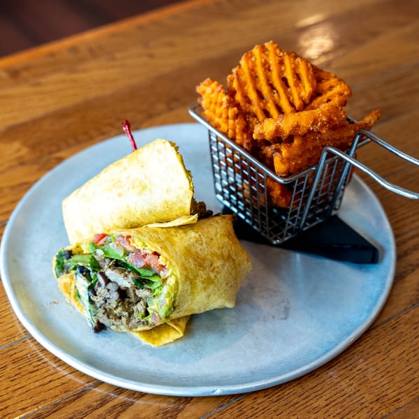 A chicken and avocado wrap with a side of waffle fries in a metal basket on a wooden table.