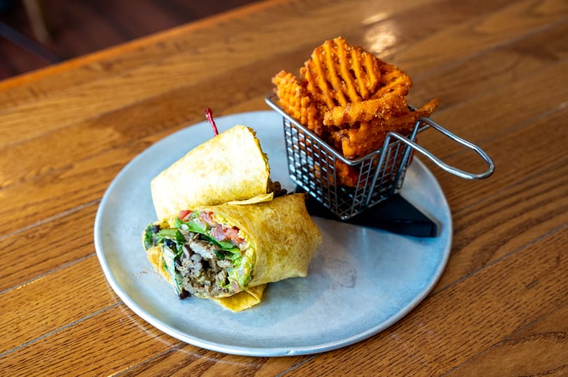 A chicken and avocado wrap with a side of waffle fries in a metal basket on a wooden table.